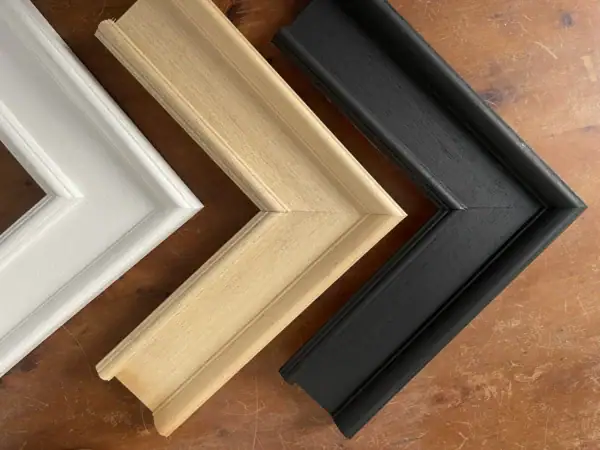 Wooden picture frame samples displayed on a table, showing natural wood, white, and black frame finishes with clean modern profiles