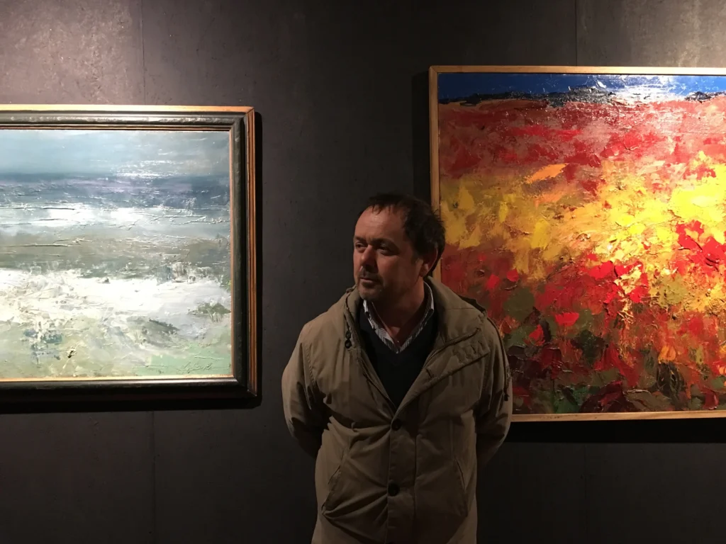 Agostino Veroni photographed during the exhibition “Il Labirinto dell’Ipnotista” at Palazzo Gallio, Lake Como, 2016, standing between two modern oil paintings.