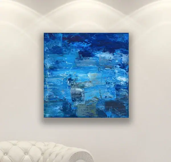The square modern abstract blue oil painting displayed in a fine black frame on a warm neutral wall above a classic white tufted sofa.