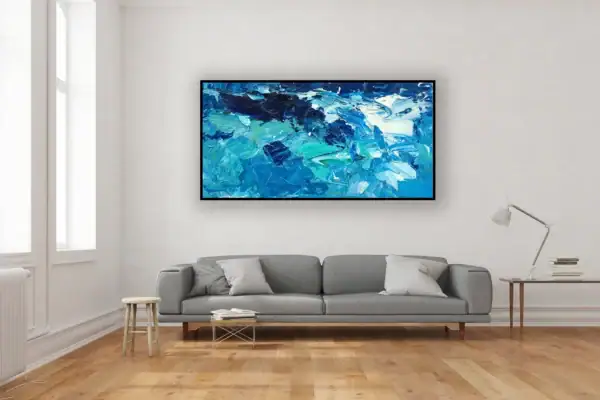 A large horizontal abstract ocean oil painting with blue waves hanging on a white wall in a modern living room with a grey sofa.