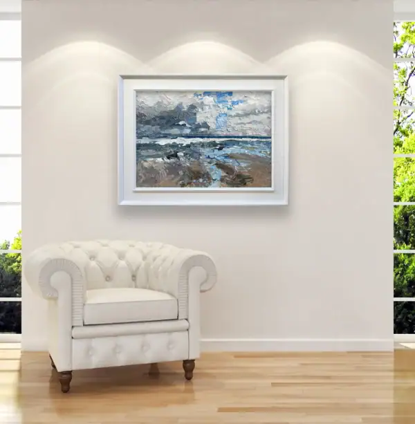 The horizontal coastal landscape oil painting framed in white on a contemporary living room wall next to an elegant leather armchair.