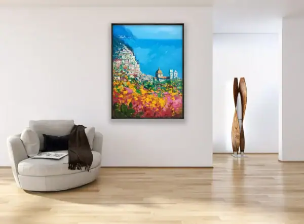 A large framed vertical painting of the Amalfi Coast hanging in a minimalist modern living room above a grey designer armchair, illustrating the scale and vibrant colors of the piece.