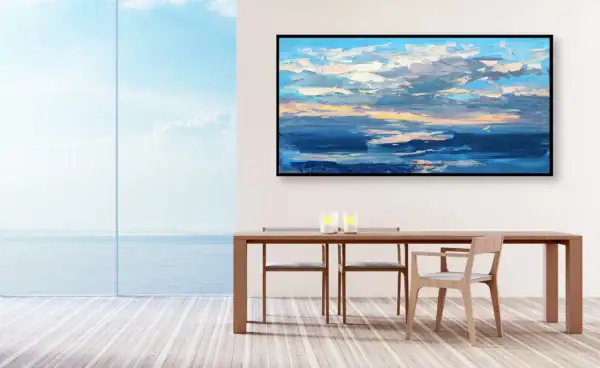 The large horizontal gold sunset seascape hanging on a minimalist white wall in a luxury dining room with wooden table and open ocean view.