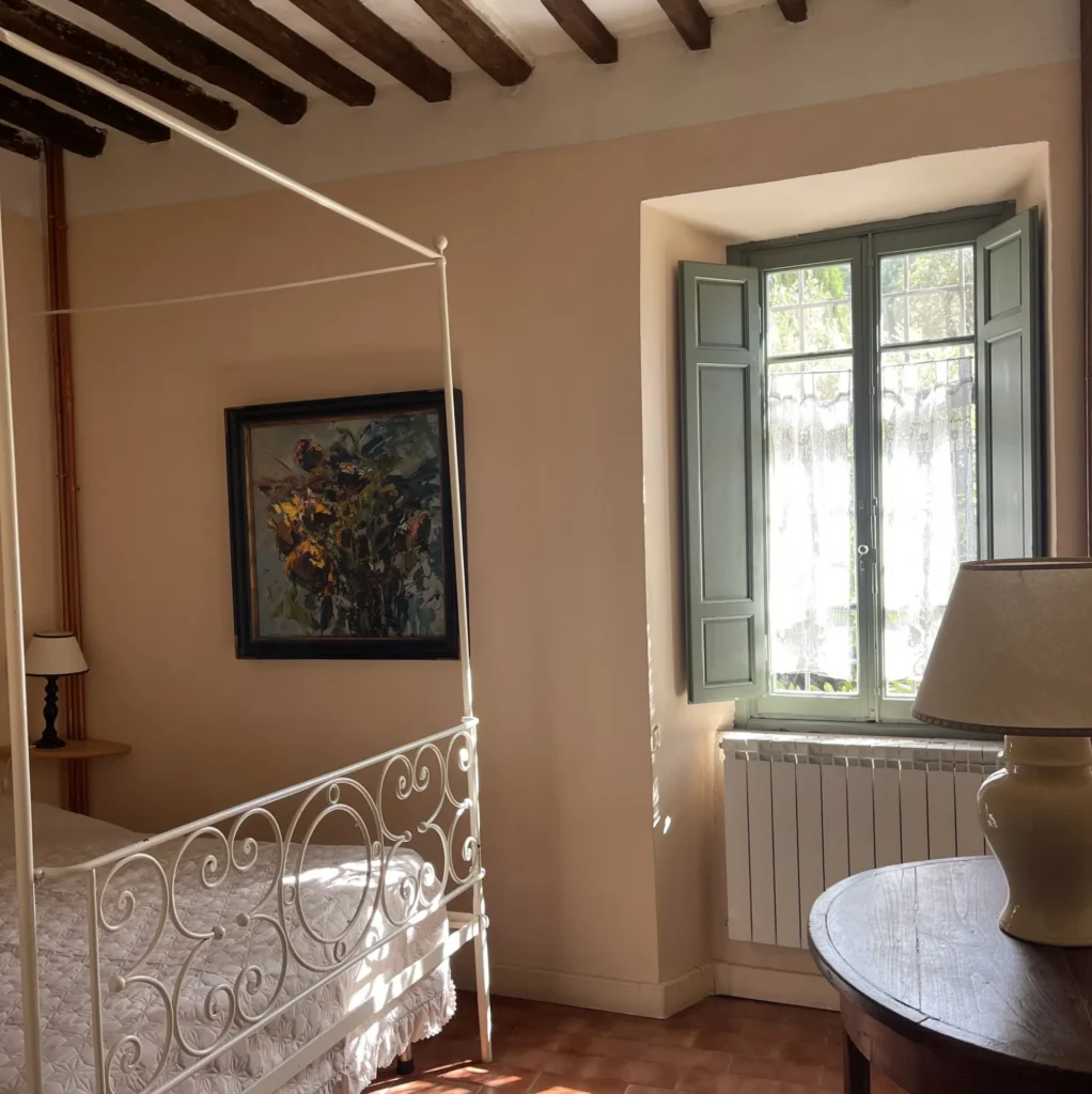 Authentic Tuscan bedroom with a white wrought iron canopy bed, wooden beam ceilings, and original fine art paintings by Agostino Veroni at Villa Minghetti holiday rental.