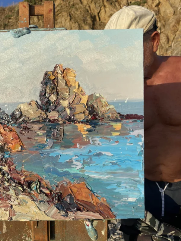 An artist during an en plein air oil painting session on the coast, working on a canvas of textured coastal rocks.