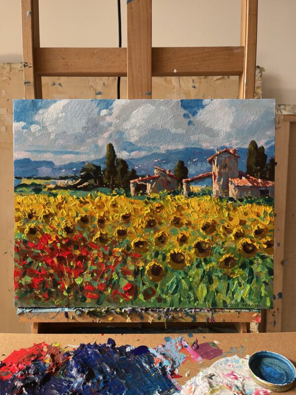 This Tuscany oil painting is displayed on a wooden studio easel alongside an artist palette. Full view photo of the original artwork resting in an art studio, featuring vibrant golden sunflowers, red poppies, and rustic stone houses under a summer sky. In the foreground, a colorful palette filled with oil paints and tubes highlights the authentic hand-painted palette knife process.