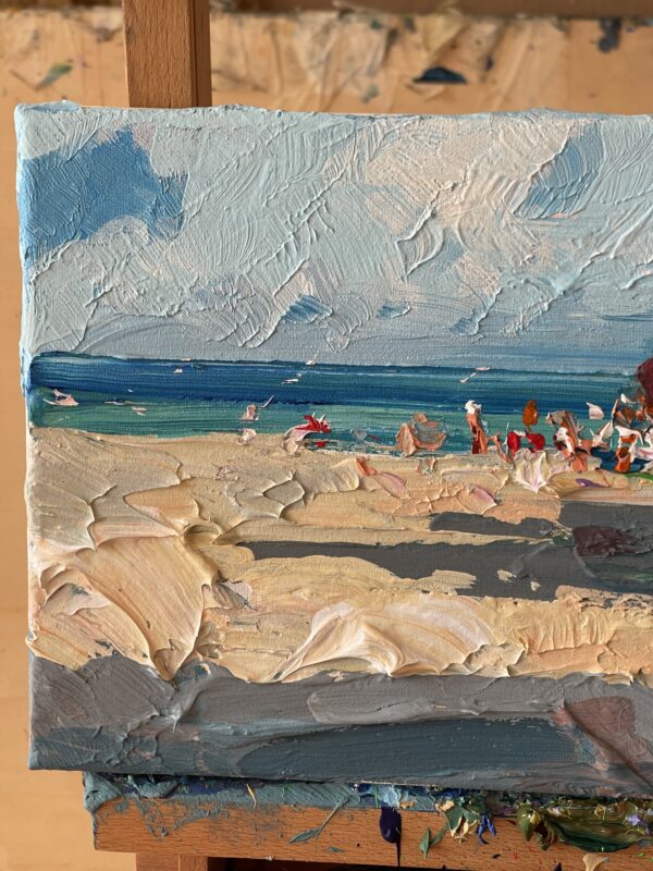 Summer beach oil painting on canvas detail on easel showing heavy impasto texture. Close-up palette knife strokes of light blue sky, sandy shore, and turquoise Mediterranean sea.