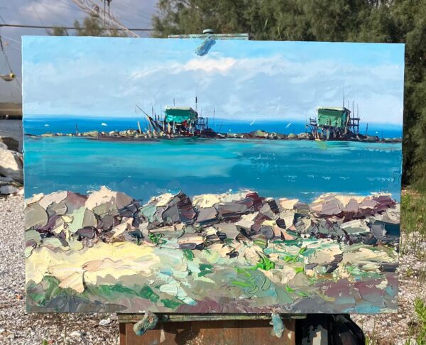 A coastal impressionist oil painting of Fishing Huts at Bocca d'Arno on an easel. Hand-painted en plein air by Agostino Veroni using a palette knife, showing rich turquoise water, green fishing cabins, and textured impasto rocks.