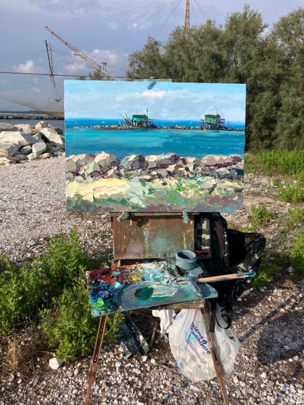 An impressionist coastal oil painting of Fishing Huts at Bocca d'Arno during an en plein air session. The canvas stands on a field easel beside a palette with oil paints, capturing vibrant Tuscan sea light and impasto textures.